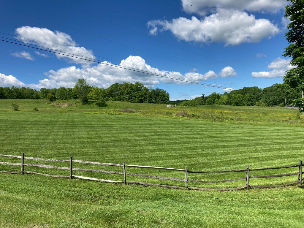A well-groomed lawn with freshly cut grass showcasing crisp, alternating stripes, set against the backdrop of Vermont farmland enclosed by rustic wood fencing. The neatly mown lawn with its distinct stripes adds a touch of elegance to the pastoral scene, while the wood fencing evokes a sense of rural charm and authenticity. The image captures the beauty of the countryside, with the manicured lawn and traditional fencing adding visual interest to the idyllic landscape.